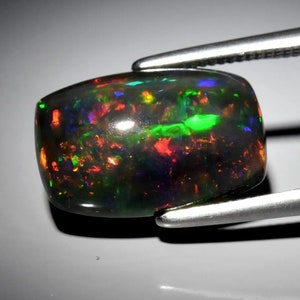 Puede incluir: Una piedra preciosa de ópalo negro con un corte cabujón. El ópalo muestra una variedad de colores, incluyendo verde, rojo y azul.