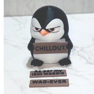 May include: A black and white penguin figurine with an orange beak and narrowed eyes, holding a brown rectangular sign that reads CHILLOUT. Two additional brown rectangular signs are placed in front, reading As per my last waddle and WAD-EVER.
