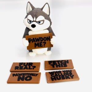 May include: A gray and white husky figurine with a textured surface, holding a brown rectangular sign with the black text PAWDON ME?. Four additional brown signs are included with the black text FUR REAL?, FETCH THIS, PAWSITIVELY NO, and WHO YOU CALLING HUSKY.