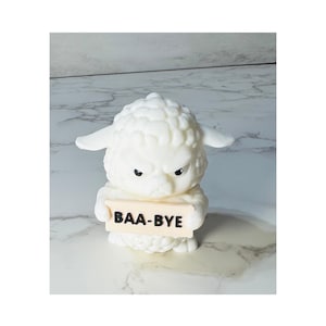 May include: A small white sheep figurine with a textured woolly appearance and a grumpy facial expression. The sheep is holding a rectangular beige sign in its front hooves that features the text BAA-BYE printed in bold black capital letters.
