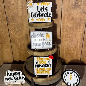 May include: A tiered wooden tray with New Year's Eve themed decorations. The decorations include signs that say "Lets Celebrate", "Cheers to the New Year", and "Kiss me NOW MINOGHT is past my BEDTIME". A clock shows the time as midnight.