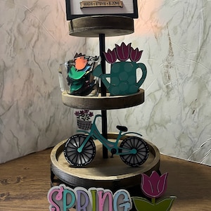 May include: A three-tiered wooden stand decorated with spring-themed items. The top tier features a sign that reads "Flower Market Open Daily Seeds Stems Blooms". The middle tier has a glass jar filled with colorful flower petals and a turquoise watering can with pink flowers. The bottom tier has a blue bicycle with a basket of flowers, a wooden sign that says "Spring", and a pink tulip.
