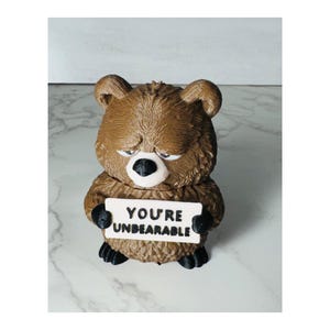 May include: A brown bear figurine with a textured fur finish, black nose and paws, and a white rectangular sign held in its hands. The sign features the black text "YOU'RE UNBEARABLE" printed in a clear, bold font. The bear has a sad expression.