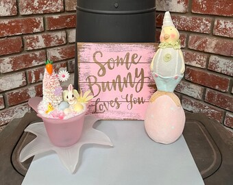 May include: A pink wooden sign with the text "Some Bunny Loves You" in brown lettering. A pink ceramic Easter egg with white polka dots and a white chick figurine with a blue and white ruffle skirt are on a white surface. A pink ceramic bowl with a white bunny figurine, a carrot, and Easter eggs is on a white surface.