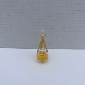 May include: A small, clear glass bottle of Dior J'adore perfume. The bottle is gold-colored and has a teardrop shape. The label on the bottle reads "J'adore" in gold lettering.