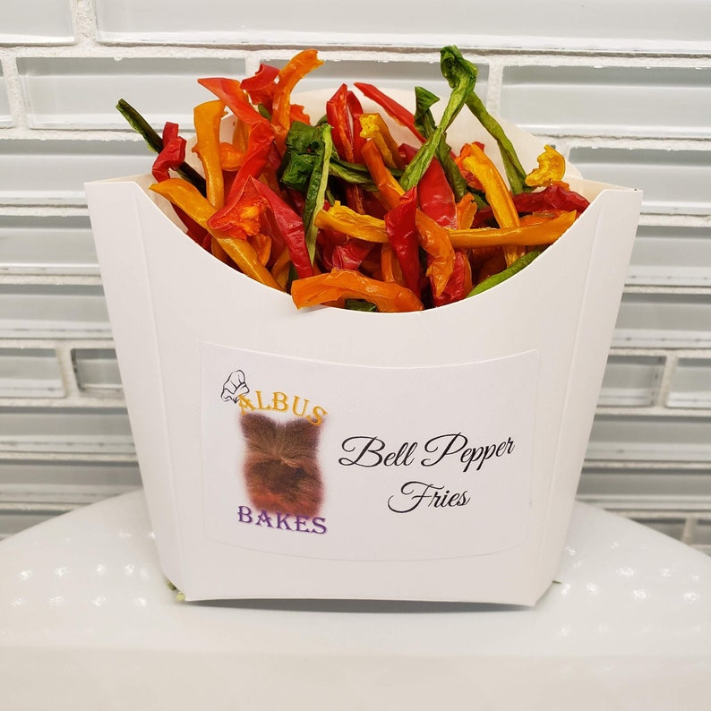 May include: A white paper container filled with colorful bell pepper fries. The container has a label that says "Albus Bakes" and "Bell Pepper Fries".