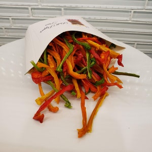 May include: A white paper bag filled with red, orange, and green dehydrated pepper strips. The bag is labeled "Bell Peppers".