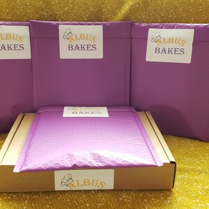 May include: Three purple padded envelopes with the text "Albus Bakes" in yellow and a chef's hat logo. A brown box with a purple padded envelope inside and a yellow label with the text "Albus Bakes" and a chef's hat logo.
