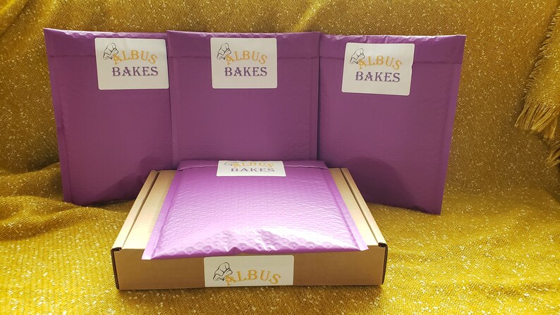 May include: Three purple padded envelopes with the text "Albus Bakes" in yellow and a chef's hat logo. A brown box with a purple padded envelope inside and a yellow label with the text "Albus Bakes" and a chef's hat logo.