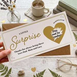 May include: A rectangular white scratch-off card with gold foil accents and a gold heart. The text reads A LITTLE Surprise, Thanks for always being there, Love Dad, Mum and Lucy, and Scratch to reveal Day out to London. The card sits on a brown envelope.