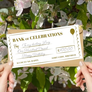May include: A novelty cheque from the Bank of Celebrations, made out to "Lucy" for £100.00, featuring a balloon graphic. The cheque is held against a backdrop of white flowers and green foliage. The text reads "Happy Birthday".