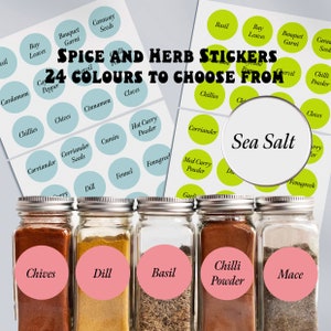 May include: A set of six spice jars with pink labels. The labels have black text and include the names: Chives, Dill, Basil, Chilli Powder, and Mace. The jars are filled with various spices and herbs. The image also includes a sheet of sticker labels with various spice and herb names.