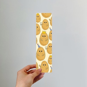May include: A yellow bookmark with a repeating pattern of cartoon potatoes wearing party hats and crowns. The potatoes are smiling and have brown eyes.