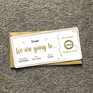 May include: A white card with gold text that says "We are going to..." and "Louise" with a gold heart symbol. The card has a gold circle with the text "Westend" that is scratched off to reveal the destination. The card also has the text "Departure 06/09/2024", "Time Two o'clock", and "Venue Lendan".