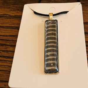 May include: A gold pendant necklace with a black and white patterned rectangular pendant. The pendant is on a black cord.