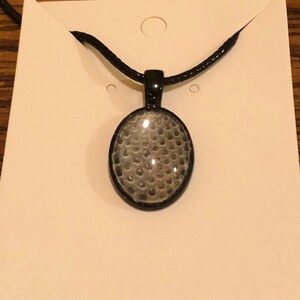 May include: Black oval pendant necklace with a textured, gray design. The pendant is set in a black frame and hangs from a black cord. The necklace is displayed on a white card.