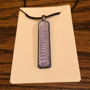 May include: A purple and white rectangular pendant with a snakeskin pattern hangs from a black cord necklace.