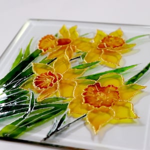 May include: A square glass artwork showcasing daffodils. The flowers have yellow petals and orange centres, outlined in gold. Green leaves and a gold cord complete the design, creating a decorative floral artwork.