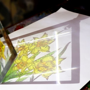 May include: A clear glass artwork depicting yellow daffodils with green leaves. The piece is set on a white surface, casting a shadow of the flowers. The glass is angled, revealing the artwork from different viewpoints. The floral design is visible.