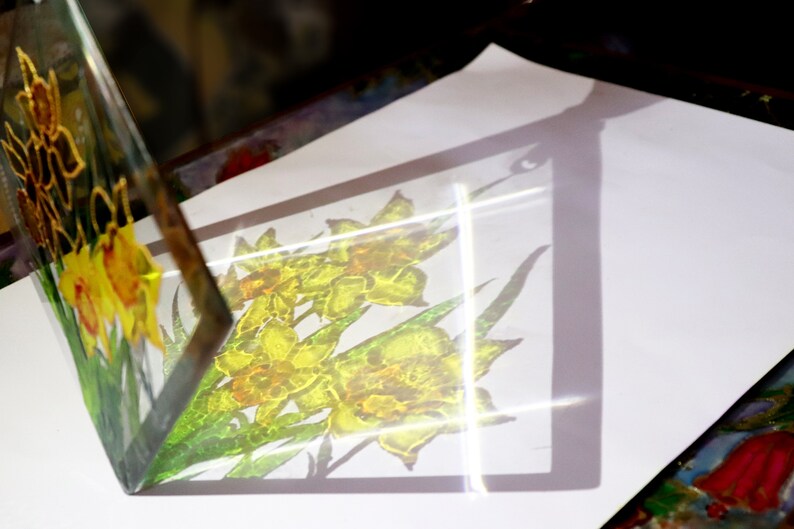 May include: A clear glass artwork depicting yellow daffodils with green leaves. The piece is set on a white surface, casting a shadow of the flowers. The glass is angled, revealing the artwork from different viewpoints. The floral design is visible.