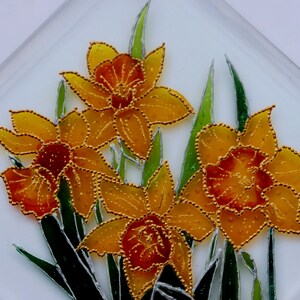 May include: A square glass decorative piece with a floral design. It features four yellow and orange daffodils with gold beaded outlines and green leaves against a white background. The daffodils are the central element of the artwork.