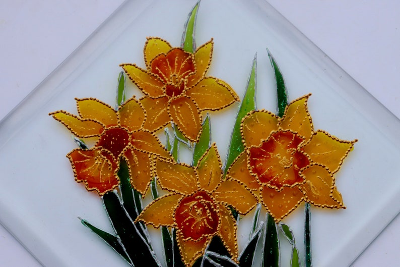 May include: A square glass decorative piece with a floral design. It features four yellow and orange daffodils with gold beaded outlines and green leaves against a white background. The daffodils are the central element of the artwork.