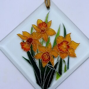 May include: A diamond-shaped glass panel with a floral design. It features yellow and orange daffodils with green leaves, outlined in gold beads. A gold ribbon is attached for hanging. The panel is suitable for decoration.