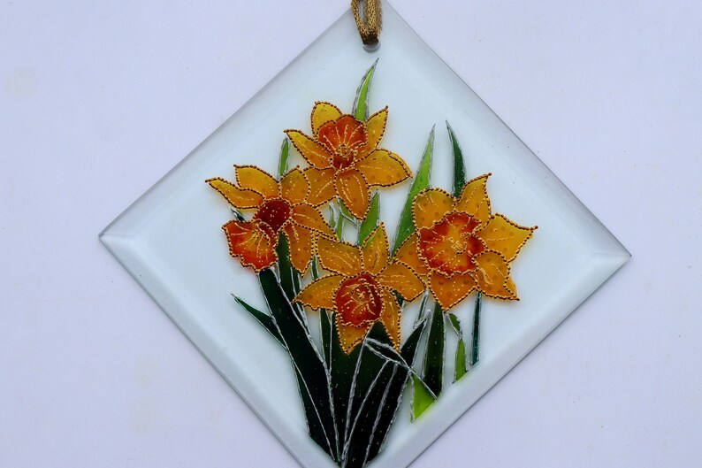 May include: A diamond-shaped glass panel with a floral design. It features yellow and orange daffodils with green leaves, outlined in gold beads. A gold ribbon is attached for hanging. The panel is suitable for decoration.
