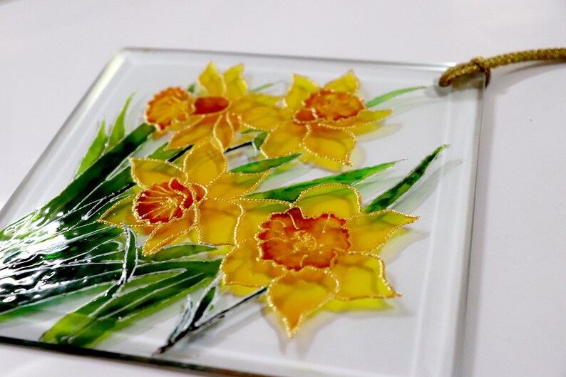 May include: A square glass artwork showcasing daffodils. The flowers have yellow petals and orange centres, outlined in gold. Green leaves and a gold cord complete the design, creating a decorative floral artwork.