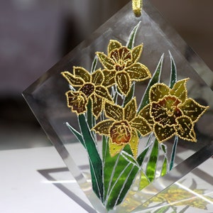 May include: A clear, diamond-shaped glass ornament with a floral design. The artwork shows yellow daffodils with green leaves and stems, outlined in gold. A gold ribbon is attached for hanging.