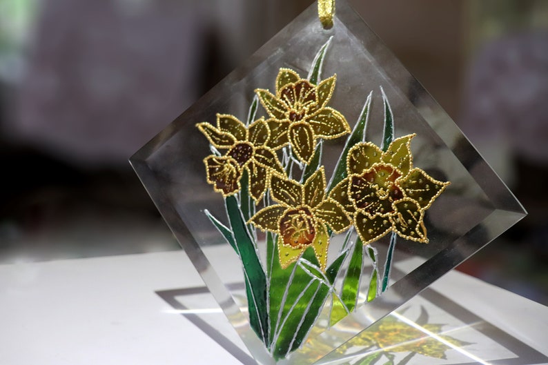 May include: A clear, diamond-shaped glass ornament with a floral design. The artwork shows yellow daffodils with green leaves and stems, outlined in gold. A gold ribbon is attached for hanging.