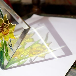 May include: A clear glass art piece depicting yellow daffodils with gold detailing and green leaves. The flowers are painted with intricate detail, and the piece is angled to show its reflection on a white surface. A decorative item.