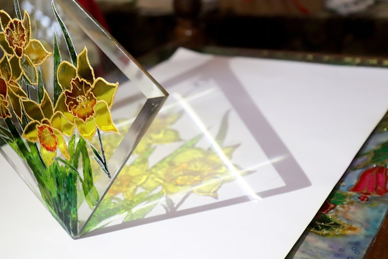 May include: A clear glass art piece depicting yellow daffodils with gold detailing and green leaves. The flowers are painted with intricate detail, and the piece is angled to show its reflection on a white surface. A decorative item.