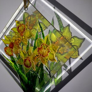 May include: A clear, diamond-shaped glass art piece featuring a bouquet of yellow and orange daffodils with green leaves. The flowers are detailed with gold accents. A gold cord is attached for hanging.