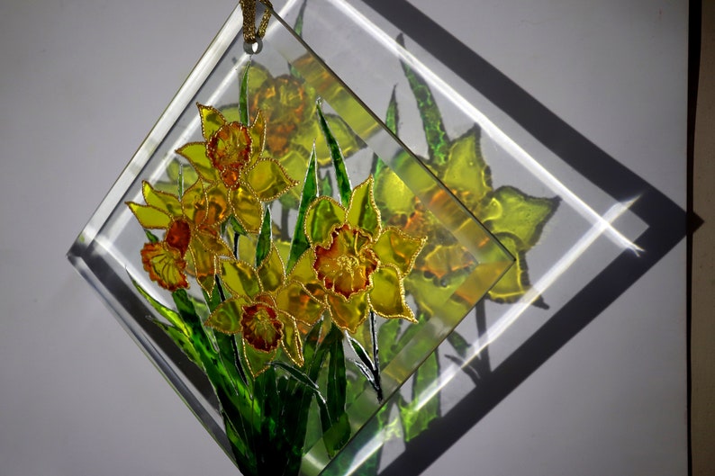 May include: A clear, diamond-shaped glass art piece featuring a bouquet of yellow and orange daffodils with green leaves. The flowers are detailed with gold accents. A gold cord is attached for hanging.