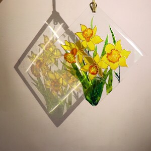 May include: A clear, diamond-shaped suncatcher with a hand-painted design of yellow daffodils and green leaves. It hangs by a gold cord, creating a shadow on the wall. The daffodils are in full bloom.