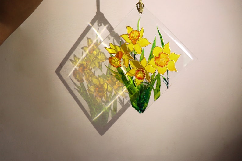 May include: A clear, diamond-shaped suncatcher with a hand-painted design of yellow daffodils and green leaves. It hangs by a gold cord, creating a shadow on the wall. The daffodils are in full bloom.