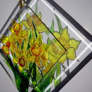 May include: A clear, diamond-shaped glass decoration featuring a bouquet of yellow daffodils with orange centres and green leaves. The flowers are outlined in gold, with a gold cord for hanging. The design is reflected in the glass.