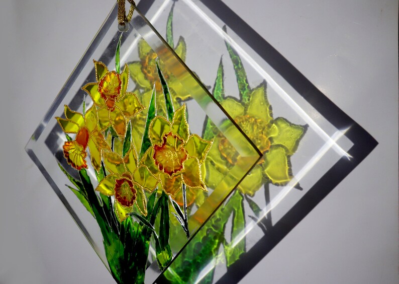May include: A clear, diamond-shaped glass decoration featuring a bouquet of yellow daffodils with orange centres and green leaves. The flowers are outlined in gold, with a gold cord for hanging. The design is reflected in the glass.