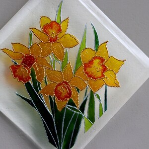 May include: A decorative glass panel with a daffodil design. The daffodils have yellow petals with orange centres and are outlined in gold. Green leaves and stems complete the floral arrangement, set against a white background.