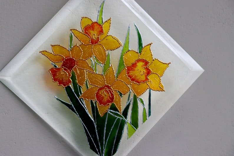 May include: A decorative glass panel with a daffodil design. The daffodils have yellow petals with orange centres and are outlined in gold. Green leaves and stems complete the floral arrangement, set against a white background.