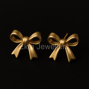 Puede incluir: Un par de pendientes de bot&oacute;n dorados con forma de lazo. Los pendientes son de metal mate y est&aacute;n sobre un fondo negro. El texto "Zk Jewelers" es visible.