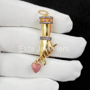 Op de afbeelding: Een goudkleurige hanger in de vorm van een hand die een roze hart vasthoudt. De hanger heeft twee banden met kleine witte en roze stenen. De tekst "Ekta Jewellers" is zichtbaar op de hanger.