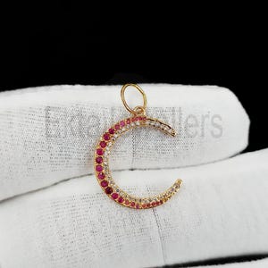 May include: A gold-colored crescent moon pendant with a loop at the top. The moon is encrusted with alternating red and clear gemstones. The pendant is displayed on a white surface against a black background.