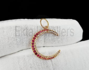 May include: A gold-colored crescent moon pendant with a loop at the top. The moon is encrusted with alternating red and clear gemstones. The pendant is displayed on a white surface against a black background.