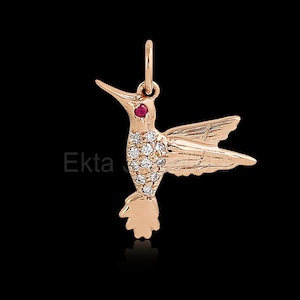 May include: A rose gold hummingbird pendant with a red gemstone eye and a body encrusted with small, clear gemstones. The wings are detailed, and the pendant has a loop for a chain. The word "Ekta" is visible on the left side.