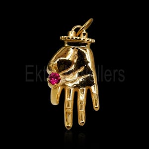 Op de afbeelding: Een goudkleurige hanger in de vorm van een hand, met een roze edelsteenring. De hanger heeft een getextureerd oppervlak en een lus aan de bovenkant voor een ketting. De achtergrond is zwart.
