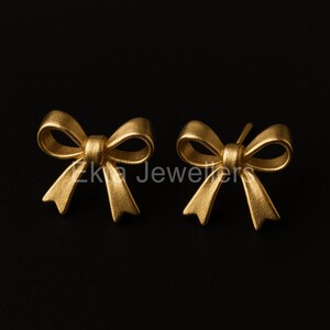 Puede incluir: Un par de pendientes de bot&oacute;n dorados con forma de lazo. Los pendientes son de metal mate y est&aacute;n sobre un fondo negro. El texto "Zk Jewelers" es visible.