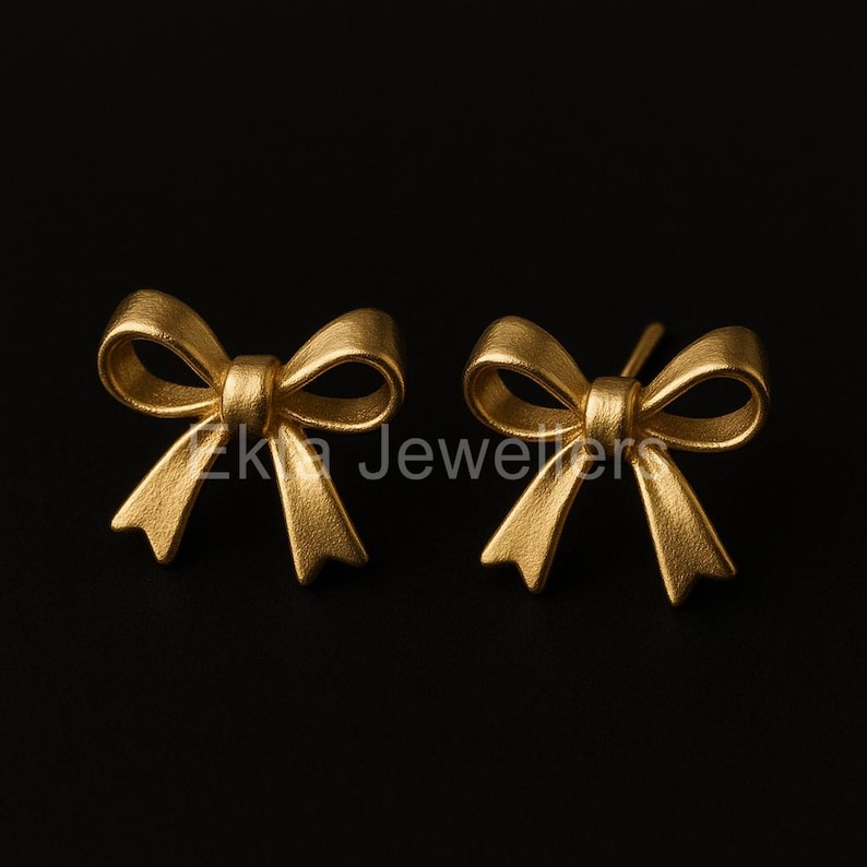 Puede incluir: Un par de pendientes de bot&oacute;n dorados con forma de lazo. Los pendientes son de metal mate y est&aacute;n sobre un fondo negro. El texto "Zk Jewelers" es visible.