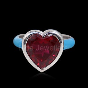 Puede incluir: Un anillo plateado con una piedra preciosa roja oscura facetada en forma de corazón. La banda es azul claro. El anillo está sobre un fondo negro. El anillo es una joya.